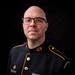 A bald male soldier wearing a U.S. Army dress uniform (black with gold piping) poses for a formal portrait against a black background. He wears glasses and displays multiple medals, ribbons, and insignia consistent with a senior non-commissioned officer rank. The uniform includes a sleeve patch reading 'U.S. Army' and sergeant first class or master sergeant chevrons on the sleeve.