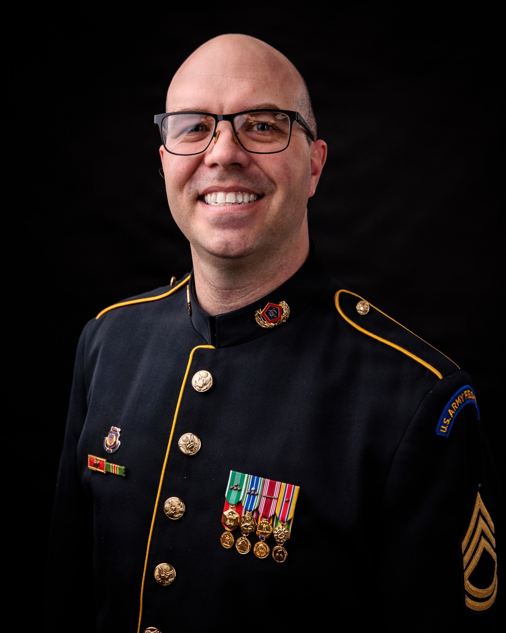 A bald man wearing glasses is photographed in a formal U.S. Army dress uniform (blue/black with gold piping) against a black background. He is smiling broadly and displays multiple service medals, ribbons, and rank insignia. The shoulder patch reads 'U.S. Army Band' and his sleeve chevrons indicate the rank of Sergeant First Class or Master Sergeant.