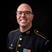 A bald man wearing glasses is photographed in a formal U.S. Army dress uniform (blue/black with gold piping) against a black background. He is smiling broadly and displays multiple service medals, ribbons, and rank insignia. The shoulder patch reads 'U.S. Army Band' and his sleeve chevrons indicate the rank of Sergeant First Class or Master Sergeant.