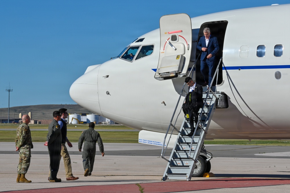 A white military or government aircraft is parked on a tarmac at what appears to be a military airfield. Several individuals, including uniformed military personnel in camouflage and flight suits, are gathered near the aircraft, while a civilian in a blue suit descends the boarding stairs. The setting suggests an official arrival or departure ceremony at a U.S. military installation.