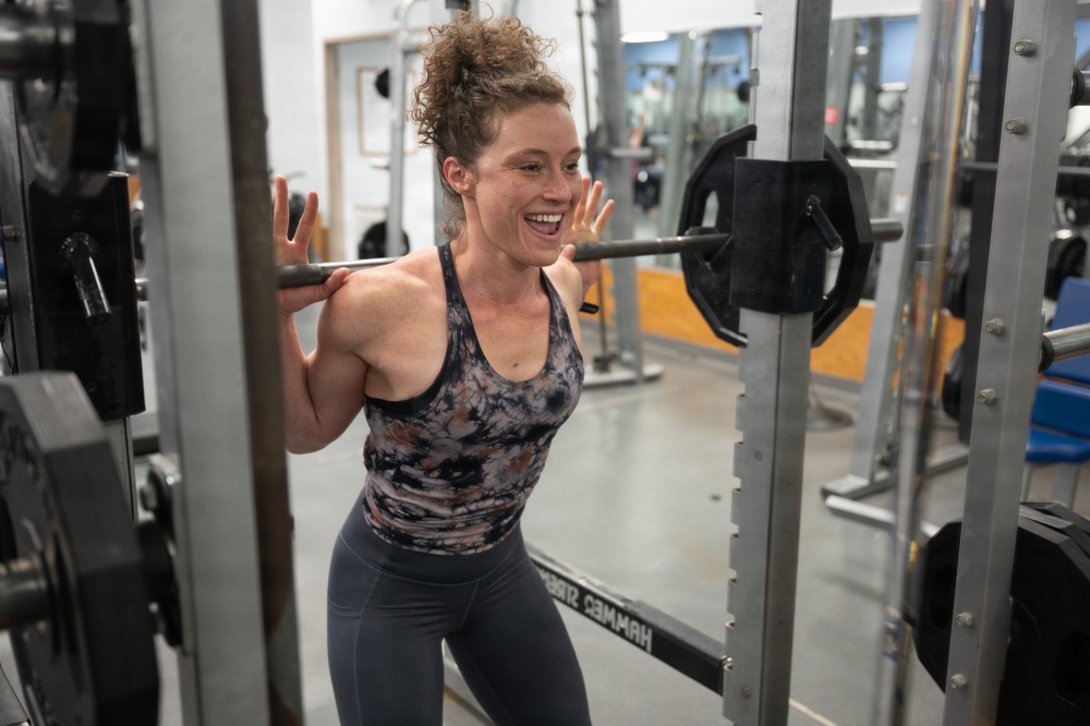 A woman is performing a barbell back squat exercise inside a gym, positioned within a power rack. She is smiling and appears energetic, with the barbell resting across her upper back/traps. The gym environment includes multiple machines, mirrors, and weight plates visible in the background.