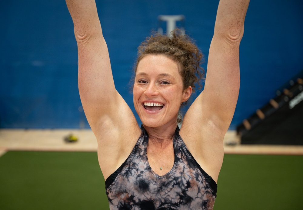 A woman is photographed in what appears to be a gym or fitness facility with her arms raised overhead, displaying a broad, joyful smile. She is wearing a tie-dye athletic tank top and has curly hair pulled up. The setting includes a blue wall, green turf flooring, and gym equipment visible in the background.