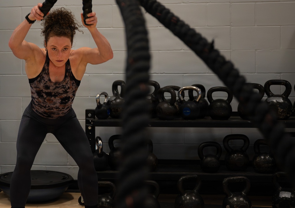 A muscular woman is performing a battle rope exercise in a gym setting, gripping two thick black ropes raised above her head in a double-arm slam position. She is wearing a patterned tank top and dark leggings, with an intense, focused expression. Behind her is a well-stocked kettlebell rack mounted against a cinder block wall.