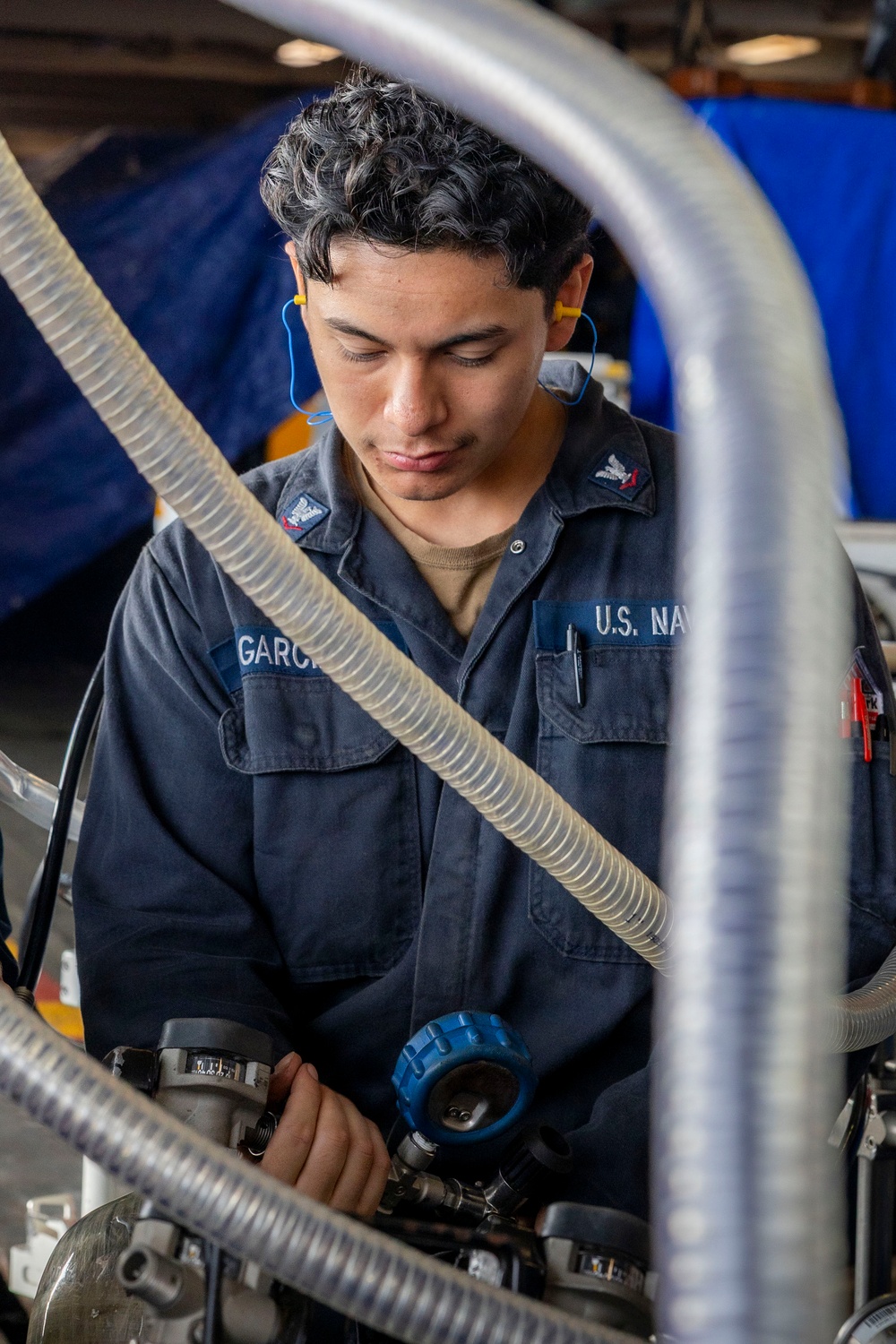 A U.S. Navy sailor with the last name Garcia is performing maintenance or technical work on mechanical equipment in what appears to be a hangar or maintenance bay. The sailor is wearing standard Navy working uniform (NWU) with rank insignia and has yellow foam earplugs inserted for hearing protection. The image is framed through metal tubing and flexible hoses, giving a layered industrial perspective.
