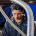 A U.S. Navy sailor with the last name Garcia is performing maintenance or technical work on mechanical equipment in what appears to be a hangar or maintenance bay. The sailor is wearing standard Navy working uniform (NWU) with rank insignia and has yellow foam earplugs inserted for hearing protection. The image is framed through metal tubing and flexible hoses, giving a layered industrial perspective.
