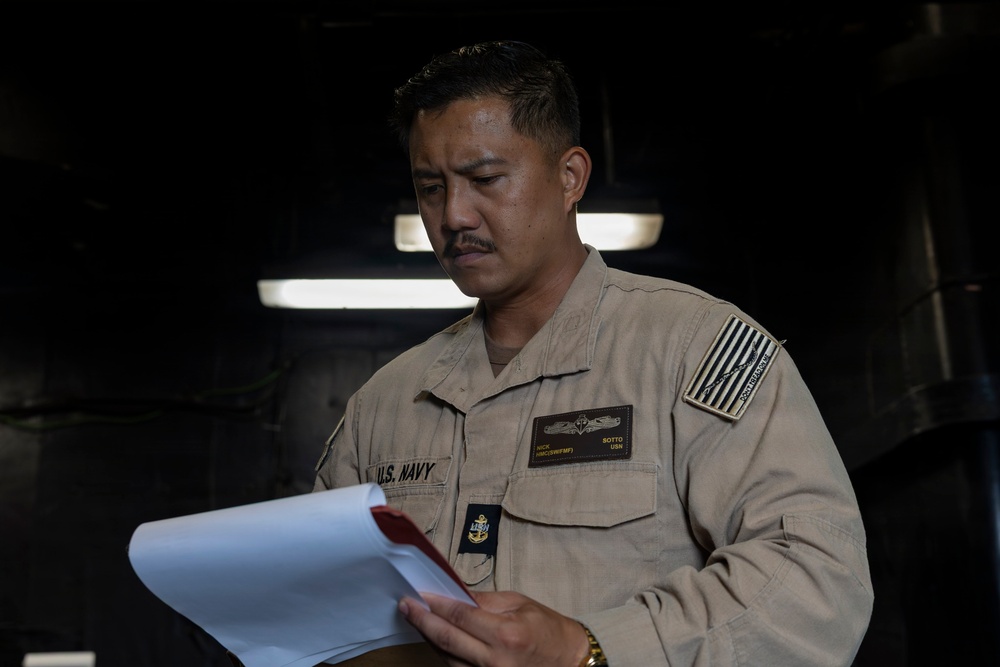A U.S. Navy sailor in tan/khaki working uniform is reviewing documents in what appears to be a shipboard interior space. The individual's name tag reads 'SOTTO' and rank insignia indicates a Chief Petty Officer (HMC) with Surface Warfare and Fleet Marine Force qualifications. The setting is dimly lit with industrial overhead lighting visible in the background.