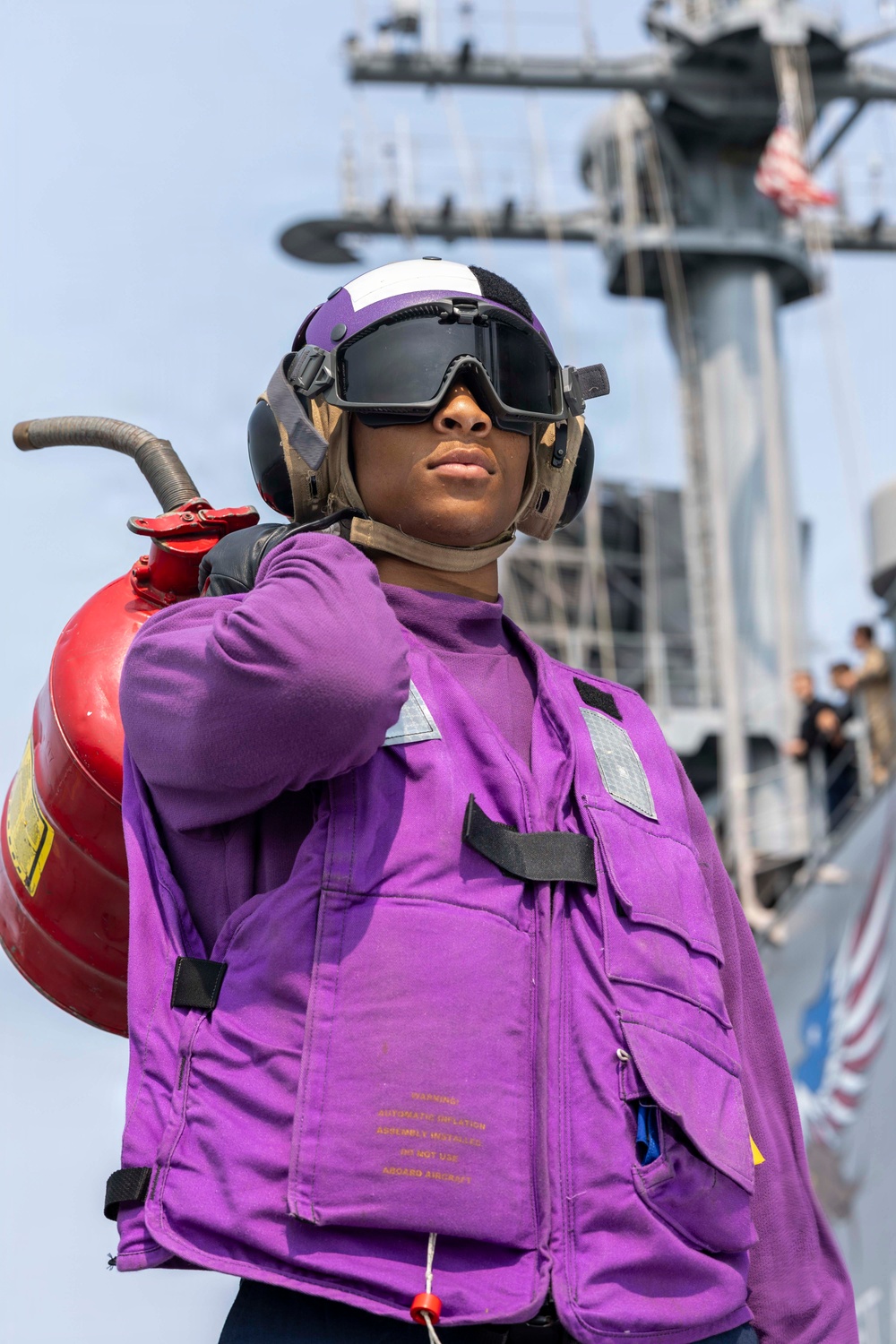 A U.S. Navy sailor wearing a purple jersey and float coat stands on what appears to be an aircraft carrier flight deck, carrying a red fire extinguisher over their shoulder. The purple color coding identifies this individual as an aviation fuels handler ('grapes') in the Navy's flight deck crew color system. A naval vessel and American flag are visible in the background.