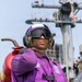 A U.S. Navy sailor wearing a purple jersey and float coat stands on what appears to be an aircraft carrier flight deck, carrying a red fire extinguisher over their shoulder. The purple color coding identifies this individual as an aviation fuels handler ('grapes') in the Navy's flight deck crew color system. A naval vessel and American flag are visible in the background.