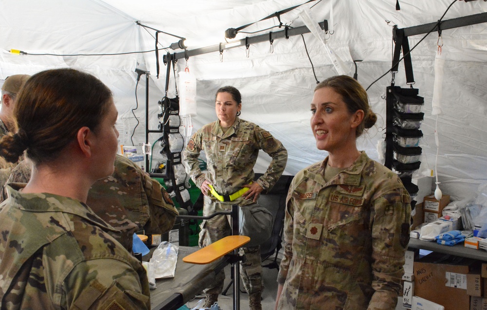 Three female U.S. Air Force personnel in OCP (Operational Camouflage Pattern) uniforms are gathered inside a military medical tent facility. The setting appears to be a field medical station or forward operating base medical unit, equipped with IV stands, medical supplies, and examination equipment. The personnel appear to be engaged in a briefing or discussion about medical operations.