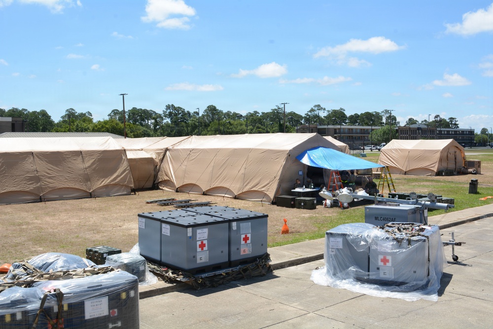 The image shows a military field medical setup, likely a forward operating base or field hospital, with large tan military tents arranged in an L-shape configuration on a grassy area. In the foreground, medical supply containers marked with Red Cross symbols are staged on pallets, along with other military equipment and supplies. The scene appears to be in the process of being assembled or prepared for operations.