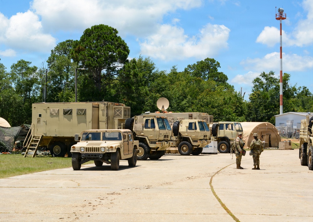 A military field operations area featuring multiple tan/desert-colored military vehicles parked on a paved surface, including a HMMWV (Humvee), tactical trucks, and a command/shelter vehicle with a satellite dish. Two uniformed soldiers are visible standing near the vehicles in what appears to be a training or staging environment. The setting includes military tents, a communications tower, and surrounding tree line consistent with a U.S. military installation.
