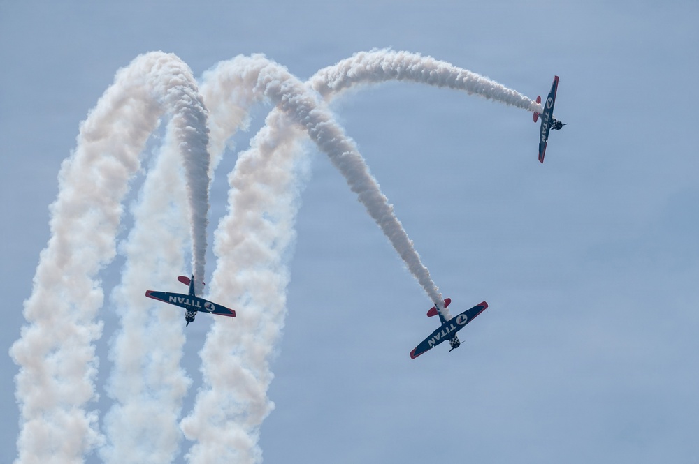 Three aerobatic aircraft branded 'TITAN' are performing a synchronized airshow maneuver against a light blue sky. The planes are producing white smoke trails that create dramatic crossing arc patterns. Two aircraft appear to be flying in close formation at lower altitude while a third diverges upward to the right.