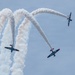 Three aerobatic aircraft branded 'TITAN' are performing a synchronized airshow maneuver against a light blue sky. The planes are producing white smoke trails that create dramatic crossing arc patterns. Two aircraft appear to be flying in close formation at lower altitude while a third diverges upward to the right.