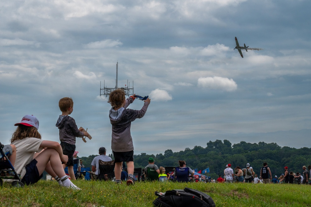 An outdoor airshow event where spectators of various ages are gathered on a grassy field watching a propeller aircraft fly overhead. Children in the foreground are holding toy model airplanes, mimicking the real aircraft in the sky. The scene captures an engaging, family-friendly aviation event under a partly cloudy sky.