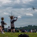 An outdoor airshow event where spectators of various ages are gathered on a grassy field watching a propeller aircraft fly overhead. Children in the foreground are holding toy model airplanes, mimicking the real aircraft in the sky. The scene captures an engaging, family-friendly aviation event under a partly cloudy sky.
