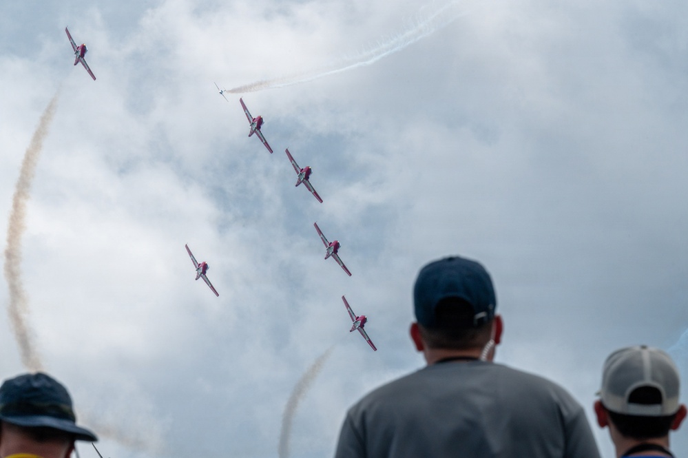 An airshow performance captured from ground level showing multiple red and white aircraft flying in formation while producing smoke trails against a partly cloudy sky. Three spectators wearing baseball caps are visible in the foreground with their backs to the camera, watching the aerial display. The aircraft appear to be executing a split or break maneuver, with one plane separating from the main group.