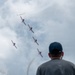 An airshow performance captured from ground level showing multiple red and white aircraft flying in formation while producing smoke trails against a partly cloudy sky. Three spectators wearing baseball caps are visible in the foreground with their backs to the camera, watching the aerial display. The aircraft appear to be executing a split or break maneuver, with one plane separating from the main group.