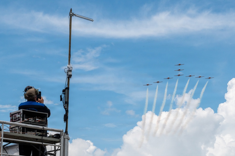 An airshow technician or controller sits atop an elevated platform equipped with communications equipment and a Yagi antenna, observing and photographing a formation of aerobatic aircraft flying in a diagonal echelon formation. The aircraft, likely a precision aerobatic team, are producing white smoke trails as they fly against a partly cloudy blue sky. The scene captures both the ground support infrastructure and the aerial performance simultaneously.