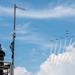 An airshow technician or controller sits atop an elevated platform equipped with communications equipment and a Yagi antenna, observing and photographing a formation of aerobatic aircraft flying in a diagonal echelon formation. The aircraft, likely a precision aerobatic team, are producing white smoke trails as they fly against a partly cloudy blue sky. The scene captures both the ground support infrastructure and the aerial performance simultaneously.