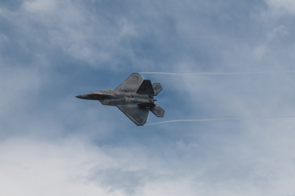 A fifth-generation stealth fighter jet, identifiable as an F-22 Raptor, is captured in flight against a partly cloudy blue sky. The aircraft is banked at a steep angle, with visible condensation vapor trails streaming from its wingtips, suggesting high-speed maneuvering. The image appears to have been taken at an airshow or military demonstration.