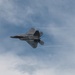 A fifth-generation stealth fighter jet, identifiable as an F-22 Raptor, is captured in flight against a partly cloudy blue sky. The aircraft is banked at a steep angle, with visible condensation vapor trails streaming from its wingtips, suggesting high-speed maneuvering. The image appears to have been taken at an airshow or military demonstration.