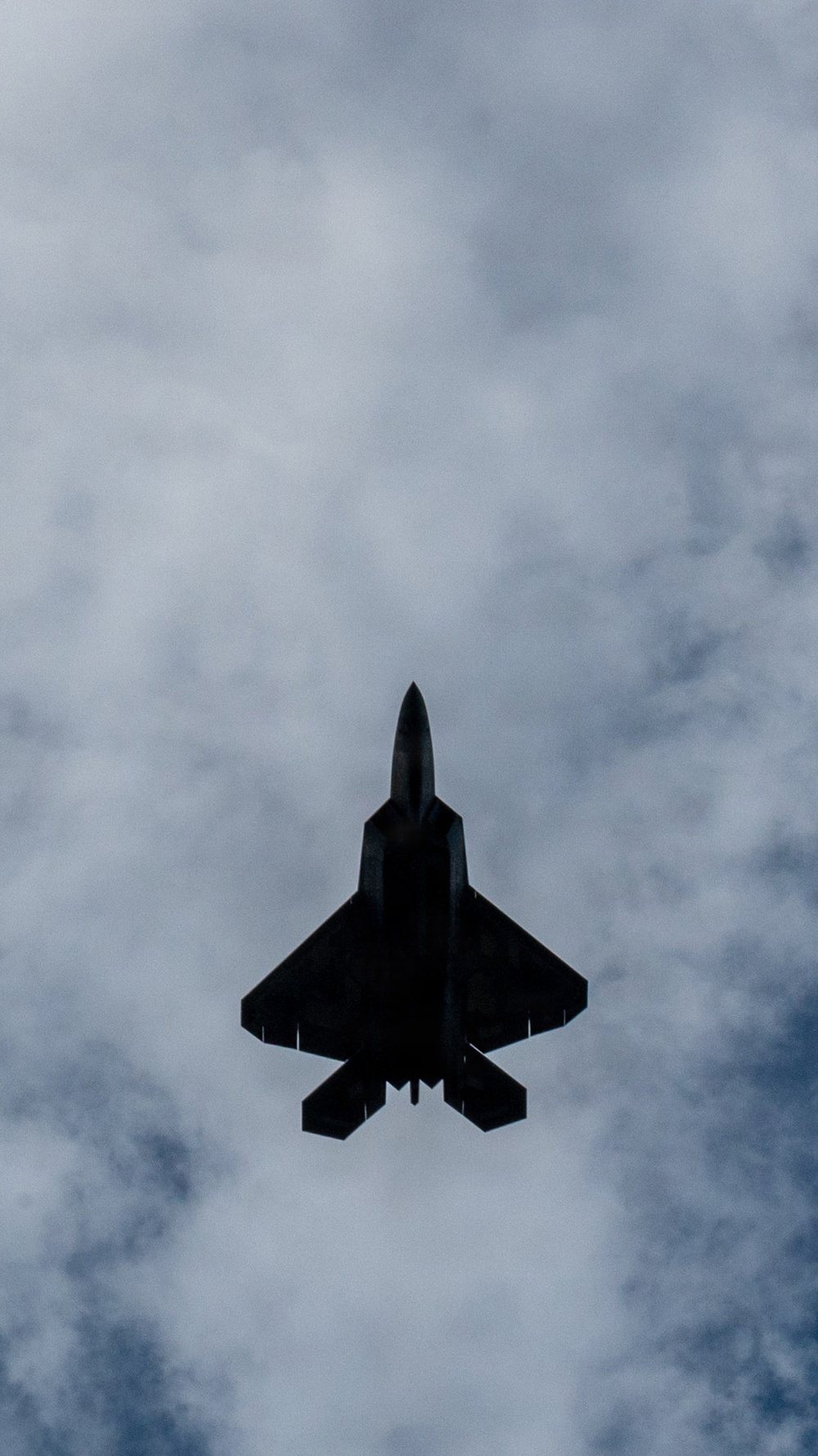 A military fighter jet is photographed from directly below against an overcast, cloudy sky. The aircraft appears as a dark silhouette, revealing its underside planform with swept delta wings, twin tail fins, and engine nozzles. The angular, stealthy design is consistent with a fifth-generation fighter aircraft such as an F-22 Raptor or F-35.