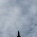 A military fighter jet is photographed from directly below against an overcast, cloudy sky. The aircraft appears as a dark silhouette, revealing its underside planform with swept delta wings, twin tail fins, and engine nozzles. The angular, stealthy design is consistent with a fifth-generation fighter aircraft such as an F-22 Raptor or F-35.