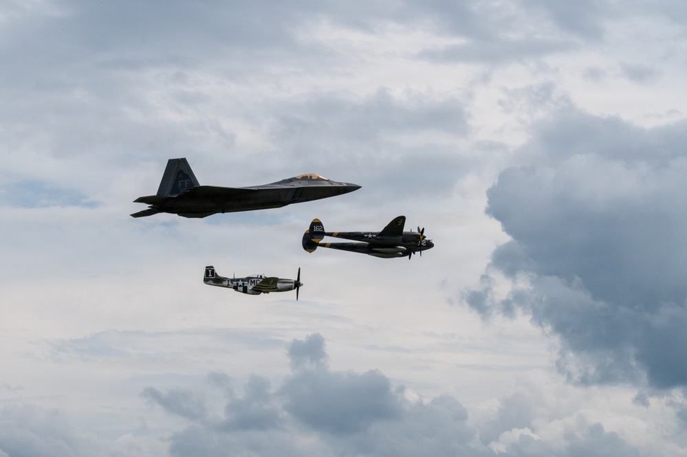 Three military aircraft are flying in formation against a partly cloudy sky, representing multiple generations of American air power. The formation includes a modern stealth fighter, a World War II-era twin-engine aircraft, and a WWII-era single-engine fighter. This appears to be a heritage flight demonstration, commonly performed at air shows.