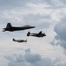 Three military aircraft are flying in formation against a partly cloudy sky, representing multiple generations of American air power. The formation includes a modern stealth fighter, a World War II-era twin-engine aircraft, and a WWII-era single-engine fighter. This appears to be a heritage flight demonstration, commonly performed at air shows.