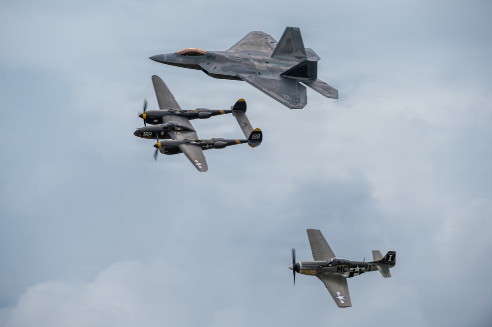 Three military aircraft from different eras fly in formation against an overcast sky, representing a 'Heritage Flight' display. The formation includes a modern stealth fighter at top, a World War II twin-boom fighter in the middle, and a WWII single-engine fighter at bottom. This type of formation is commonly performed at airshows to honor aviation history.