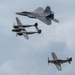 Three military aircraft from different eras fly in formation against an overcast sky, representing a 'Heritage Flight' display. The formation includes a modern stealth fighter at top, a World War II twin-boom fighter in the middle, and a WWII single-engine fighter at bottom. This type of formation is commonly performed at airshows to honor aviation history.