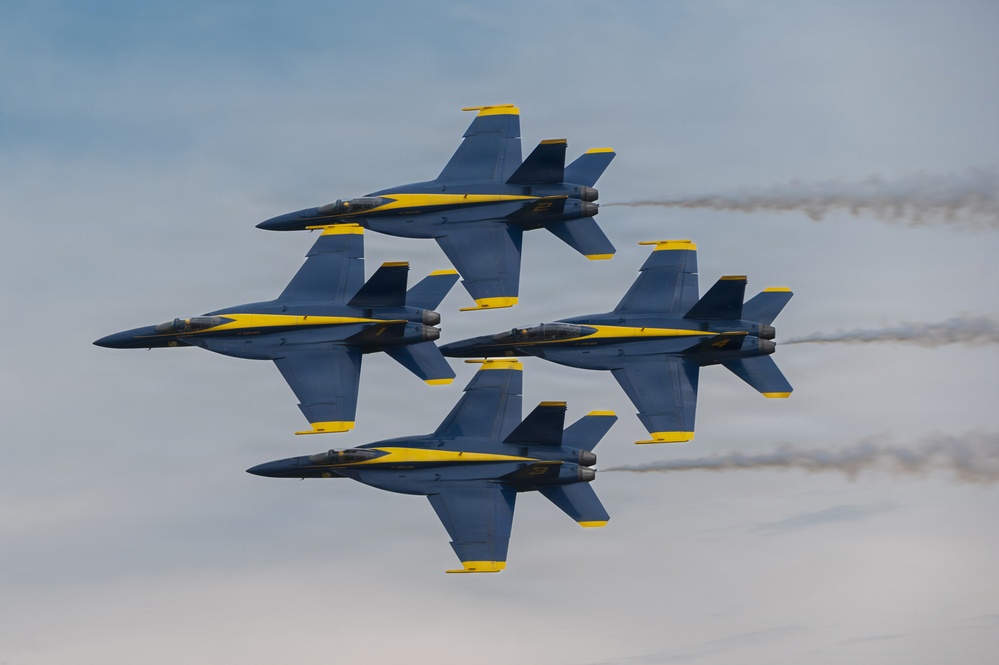 Four U.S. Navy Blue Angels aircraft are flying in a tight diamond formation against a partly cloudy sky. The jets, identifiable by their distinctive blue and yellow livery, are F/A-18 Hornets or Super Hornets performing a precision aerobatic maneuver. White smoke trails are visible streaming from the rear of the aircraft.