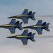Four U.S. Navy Blue Angels aircraft are flying in a tight diamond formation against a partly cloudy sky. The jets, identifiable by their distinctive blue and yellow livery, are F/A-18 Hornets or Super Hornets performing a precision aerobatic maneuver. White smoke trails are visible streaming from the rear of the aircraft.