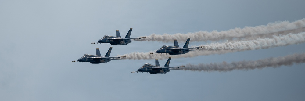 Four military jet aircraft fly in a tight diamond formation against a hazy blue-gray sky, each trailing a white smoke stream. The jets appear to be part of a precision aerobatic demonstration team, likely the U.S. Navy Blue Angels based on their blue and yellow color scheme. The formation is captured in a wide panoramic shot emphasizing the symmetry and discipline of the flight pattern.