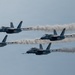 Four military jet aircraft fly in a tight diamond formation against a hazy blue-gray sky, each trailing a white smoke stream. The jets appear to be part of a precision aerobatic demonstration team, likely the U.S. Navy Blue Angels based on their blue and yellow color scheme. The formation is captured in a wide panoramic shot emphasizing the symmetry and discipline of the flight pattern.