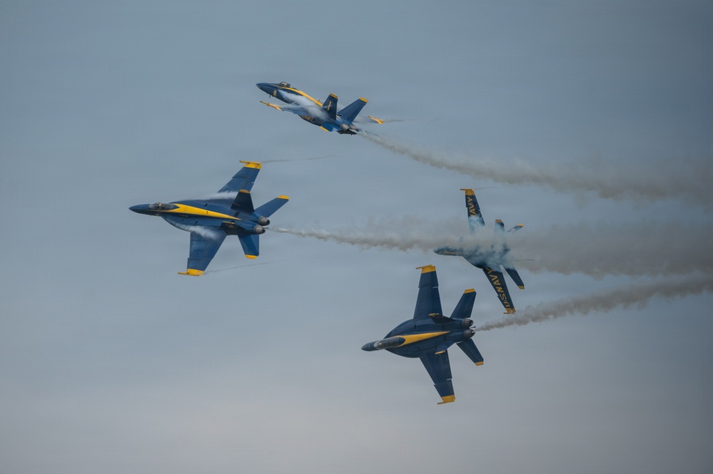 Four U.S. Navy Blue Angels F/A-18 Hornet jets are performing an aerobatic maneuver during an airshow, flying in a dynamic break formation. The aircraft are painted in their iconic blue and yellow livery with 'NAVY' markings visible on the fuselages. White smoke trails emanate from the jets, highlighting their flight paths as they diverge in multiple directions.
