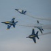 Four U.S. Navy Blue Angels F/A-18 Hornet jets are performing an aerobatic maneuver during an airshow, flying in a dynamic break formation. The aircraft are painted in their iconic blue and yellow livery with 'NAVY' markings visible on the fuselages. White smoke trails emanate from the jets, highlighting their flight paths as they diverge in multiple directions.