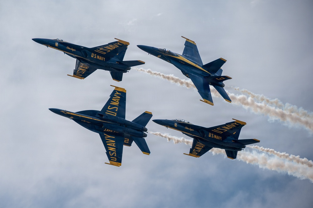Four U.S. Navy Blue Angels aircraft are performing a close formation flight maneuver at an airshow. The jets, painted in their iconic blue and gold livery, are flying in a tight diamond-like formation with two aircraft producing visible white smoke trails. The overcast sky provides a dramatic backdrop for the precision aerobatic display.