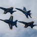 Four U.S. Navy Blue Angels aircraft are performing a close formation flight maneuver at an airshow. The jets, painted in their iconic blue and gold livery, are flying in a tight diamond-like formation with two aircraft producing visible white smoke trails. The overcast sky provides a dramatic backdrop for the precision aerobatic display.