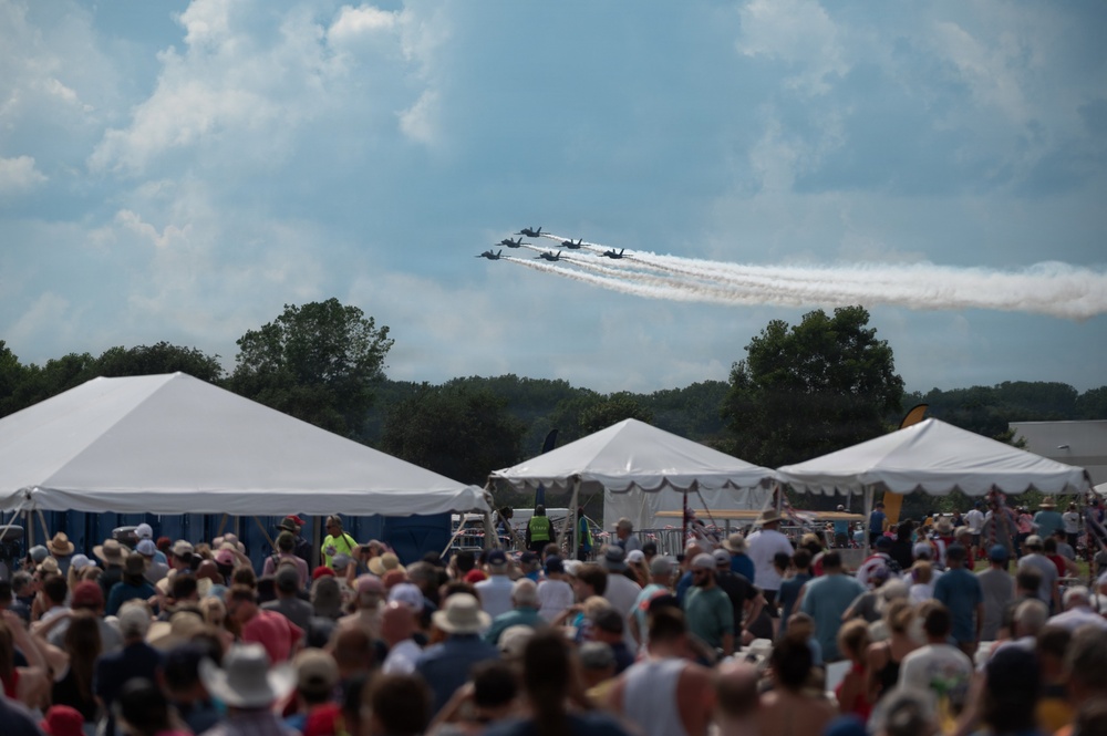 An outdoor airshow event featuring a formation of jet aircraft trailing white smoke trails across a partly cloudy sky. A large crowd of spectators watches from below, gathered near white event tents in what appears to be a fairground or airfield setting. The aircraft formation is consistent in appearance with a precision aerobatic demonstration team.