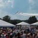 An outdoor airshow event featuring a formation of jet aircraft trailing white smoke trails across a partly cloudy sky. A large crowd of spectators watches from below, gathered near white event tents in what appears to be a fairground or airfield setting. The aircraft formation is consistent in appearance with a precision aerobatic demonstration team.