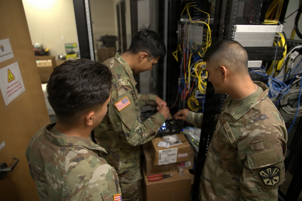 Three U.S. Army soldiers in OCP (Operational Camouflage Pattern) uniforms are working on network/communications equipment inside a server room or communications closet. They appear to be installing or troubleshooting networking hardware mounted in a rack, surrounded by numerous cables. The setting suggests a military communications or IT infrastructure maintenance operation.