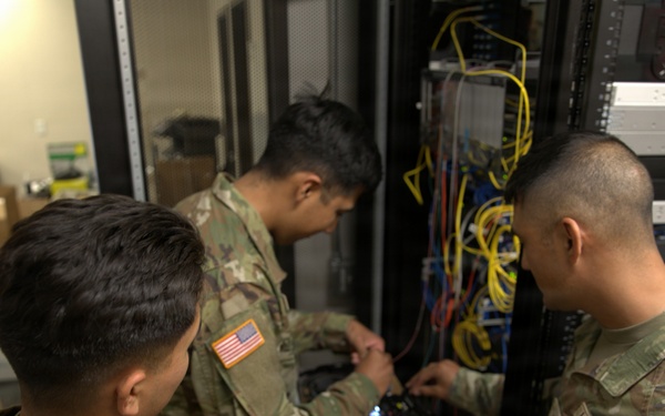 Three U.S. Army soldiers in OCP (Operational Camouflage Pattern) uniforms are working on network/communications equipment inside a server room or communications closet. They appear to be installing or troubleshooting networking hardware mounted in a rack, surrounded by numerous cables. The setting suggests a military communications or IT infrastructure maintenance operation.