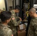 Three U.S. Army soldiers in OCP (Operational Camouflage Pattern) uniforms are working on network/communications equipment inside a server room or communications closet. They appear to be installing or troubleshooting networking hardware mounted in a rack, surrounded by numerous cables. The setting suggests a military communications or IT infrastructure maintenance operation.