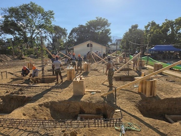 A construction site showing multiple workers actively building what appears to be a foundation structure with concrete column footings and wooden formwork. Workers wearing orange hard hats and military-style uniforms are visible, suggesting this may be a military engineering or humanitarian construction project. The site shows excavated foundation pits with rebar reinforcement and wooden bracing systems in a tropical setting.