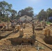 A construction site showing multiple workers actively building what appears to be a foundation structure with concrete column footings and wooden formwork. Workers wearing orange hard hats and military-style uniforms are visible, suggesting this may be a military engineering or humanitarian construction project. The site shows excavated foundation pits with rebar reinforcement and wooden bracing systems in a tropical setting.