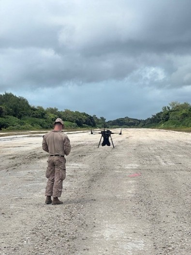 A military service member in camouflage uniform stands on a gravel or concrete strip, facing a large multi-rotor drone (UAV) positioned on the ground ahead of them. The setting appears to be a remote or expeditionary airstrip flanked by dense tropical vegetation. The scene suggests a drone pre-flight check, launch preparation, or operator training exercise.