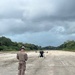 A military service member in camouflage uniform stands on a gravel or concrete strip, facing a large multi-rotor drone (UAV) positioned on the ground ahead of them. The setting appears to be a remote or expeditionary airstrip flanked by dense tropical vegetation. The scene suggests a drone pre-flight check, launch preparation, or operator training exercise.