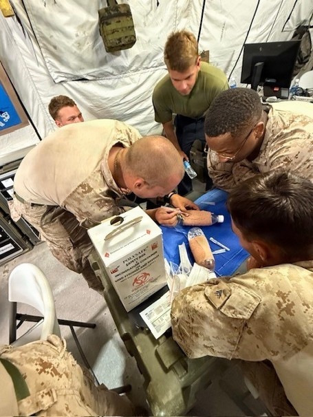 Military personnel in desert camouflage uniforms are gathered around a surface performing what appears to be a medical training exercise or procedure inside a military tent facility. A sharps disposal container labeled 'POLYSAFE SAFETY BOX' is prominently visible, along with medical supplies on a blue drape. The setting appears to be a forward operating base or field medical station.