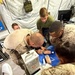 Military personnel in desert camouflage uniforms are gathered around a surface performing what appears to be a medical training exercise or procedure inside a military tent facility. A sharps disposal container labeled 'POLYSAFE SAFETY BOX' is prominently visible, along with medical supplies on a blue drape. The setting appears to be a forward operating base or field medical station.